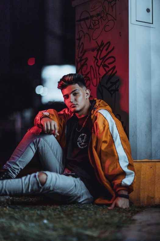 man wearing orange jacket sitting while leaning on wall